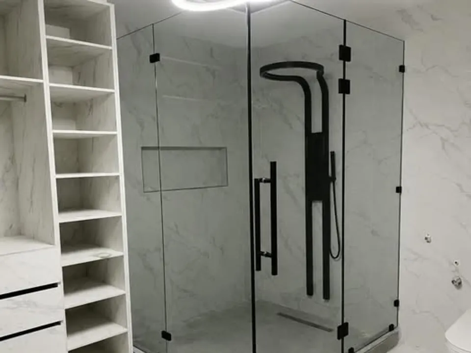 Cabina de baño en esquina de vidrio templado sin marco, con tirador y herrajes negros, en un baño de mármol
